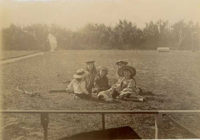 Five children seated on grass all wearing smart clothes and hats. Small dog in centre of group